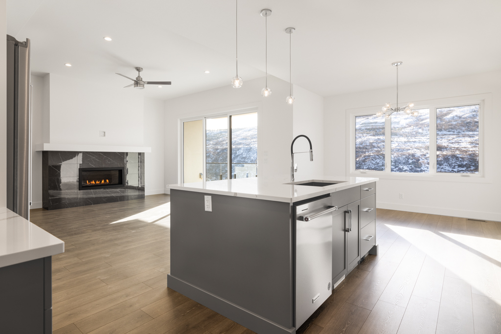 A modern kitchen with a central island, stainless steel appliances, light grey cabinetry, and recessed lighting. There's a fireplace and large windows in the adjoining living area.
