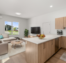 A modern open-plan kitchen and living room with light wood cabinetry, a kitchen island, an L-shaped sofa, a coffee table, and a wall-mounted TV. Virtually staged.